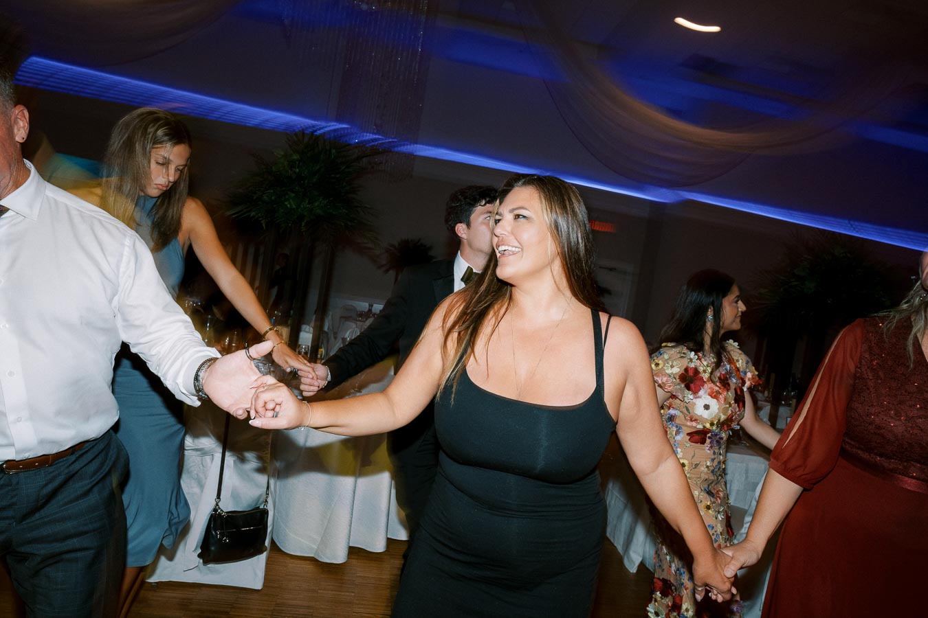 A group of people joyfully dancing in a dimly lit event hall, holding hands and smiling, creating a lively and festive atmosphere.