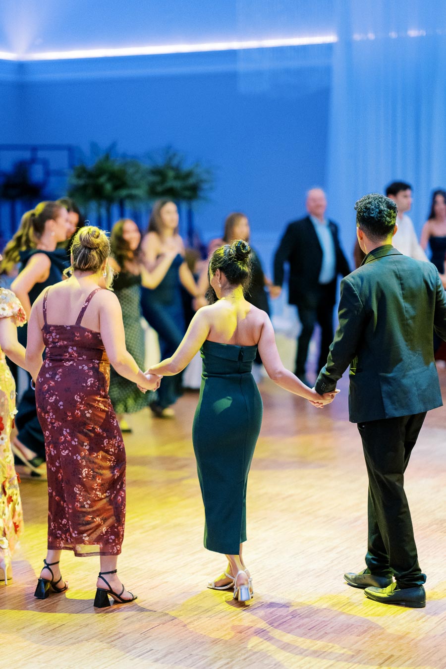 A group of elegantly dressed people dancing in a circle at a formal indoor event, with ladies in evening gowns and a man in a suit holding hands, creating a joyful and vibrant atmosphere.