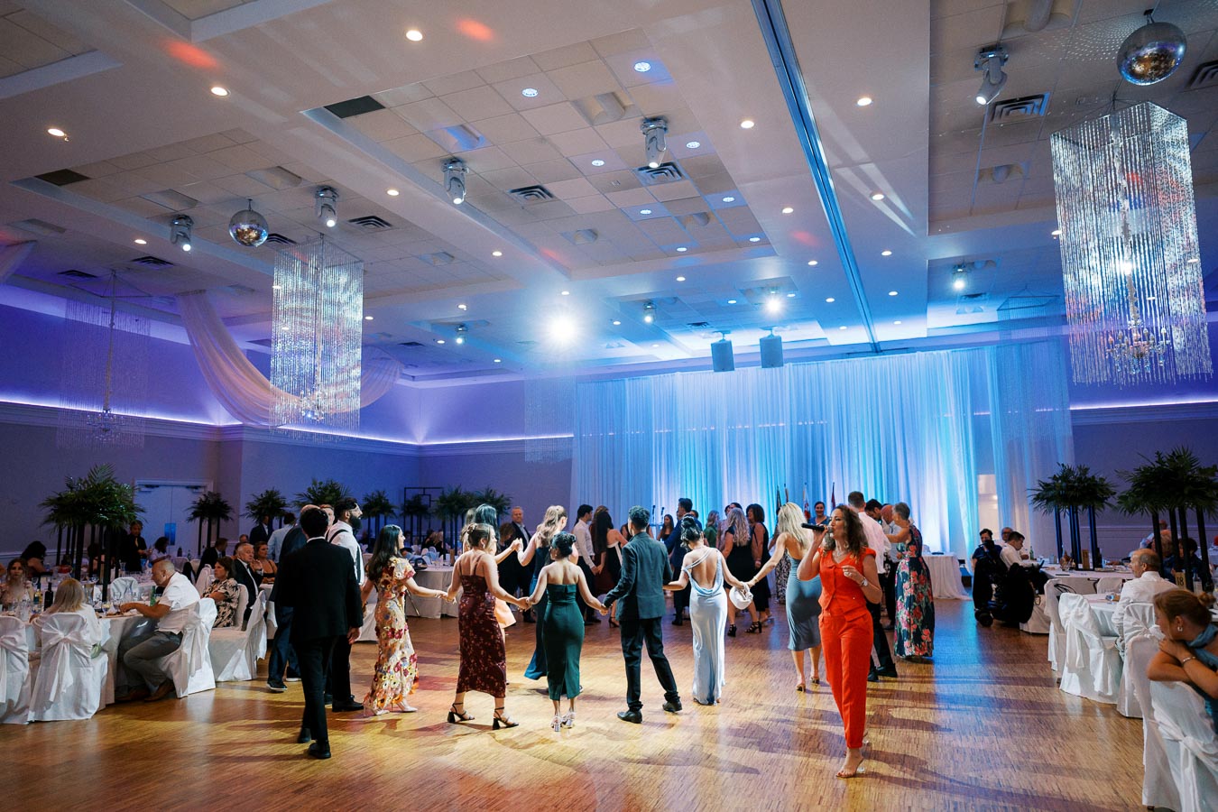 A vibrant wedding reception with elegantly dressed guests dancing in a beautifully decorated hall featuring chandeliers and ambient lighting.