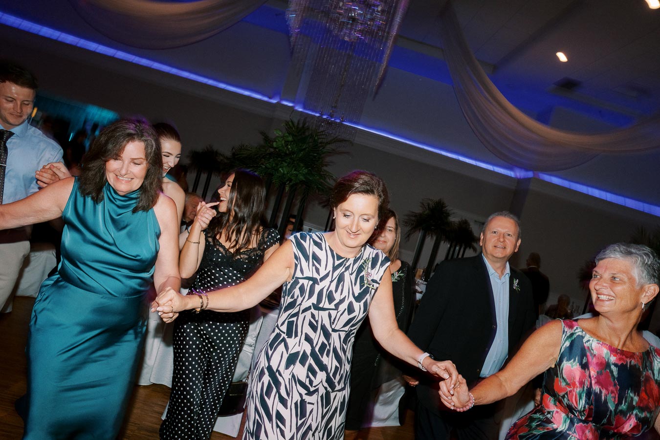 A group of people joyfully dancing in a decorated indoor venue, featuring elegant attire and colorful dresses, with blue ambient lighting and draped fabric on the ceiling.