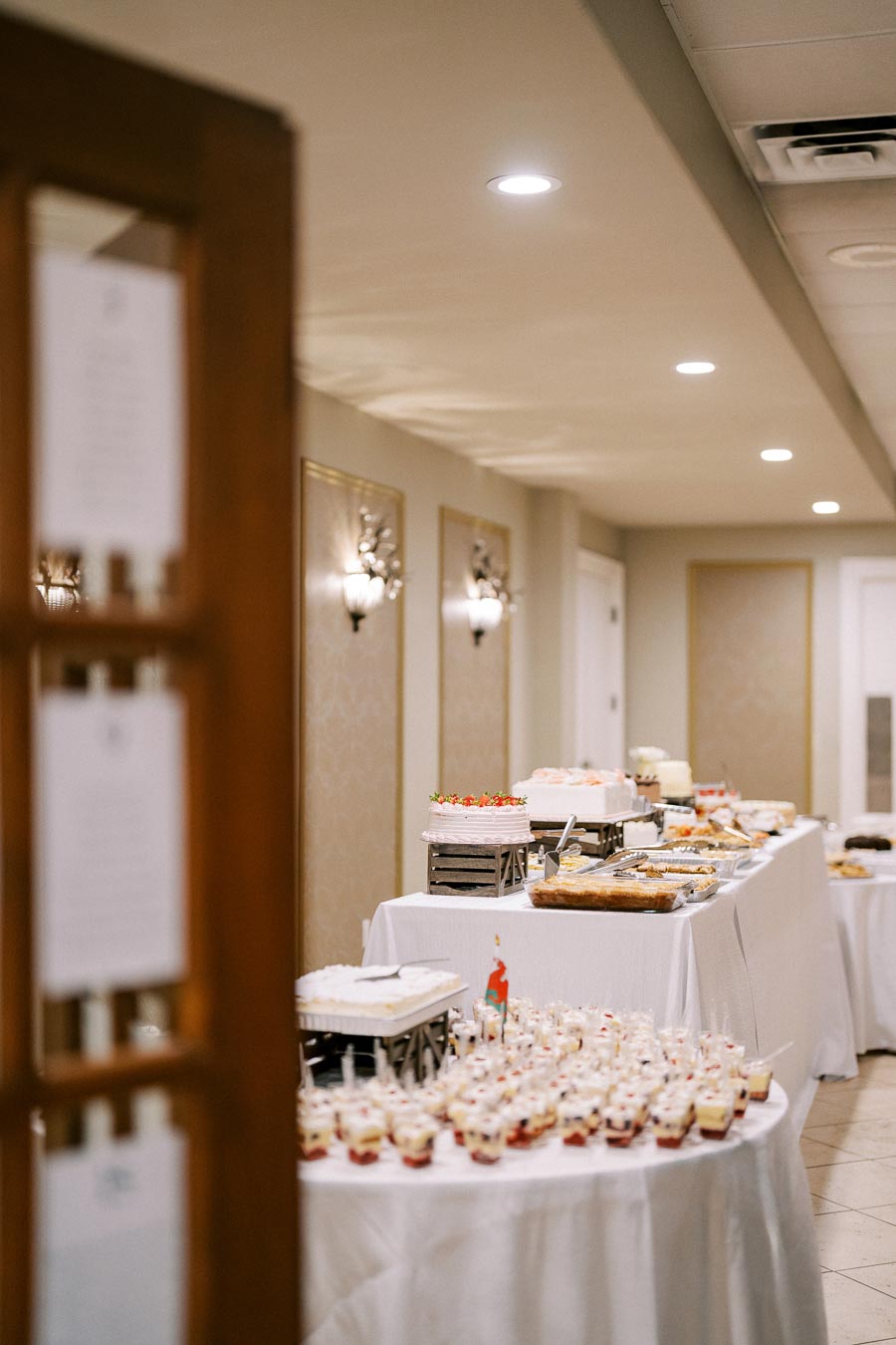 Elegant banquet hall dessert table setup with varied pastries, cakes, and desserts on white tablecloths under soft lighting, perfect for weddings and formal events.