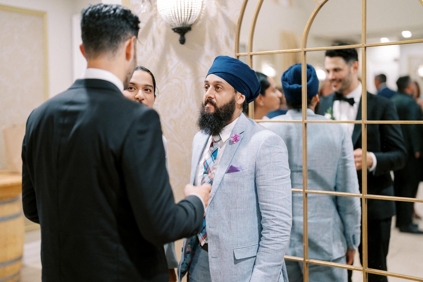 Group of well-dressed individuals engaging in conversation at a formal event, with one wearing a blue turban and light blue suit, reflected in a decorative mirror.