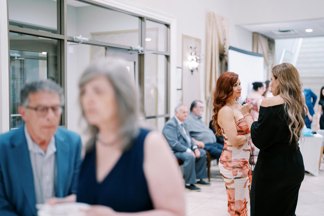 A social gathering in a modern venue with elegantly dressed attendees, featuring women conversing in the foreground and people seated in the background, suggesting a formal event atmosphere.