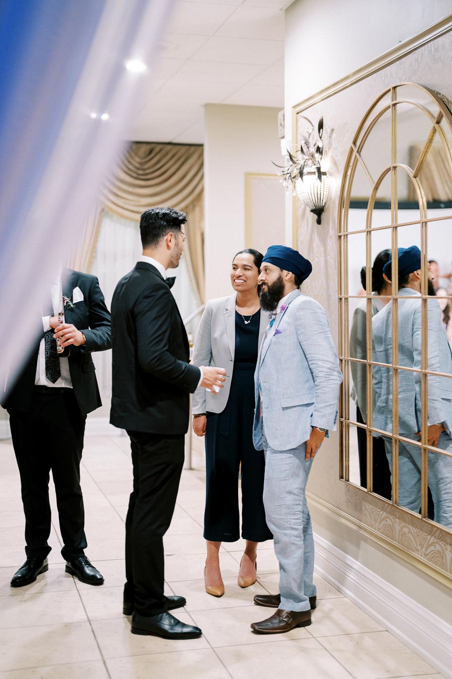 A group of well-dressed people engaged in conversation at an elegant indoor event, with mirrored wall decor and a sophisticated ambiance.