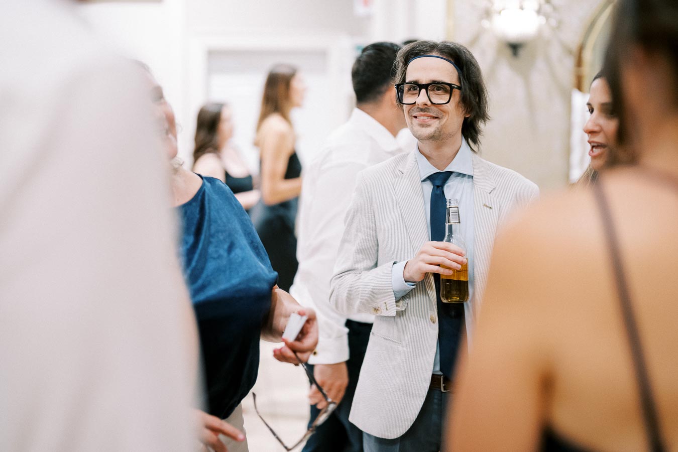 A man in glasses and formal attire holding a beer bottle at a social gathering, surrounded by people engaged in conversation.
