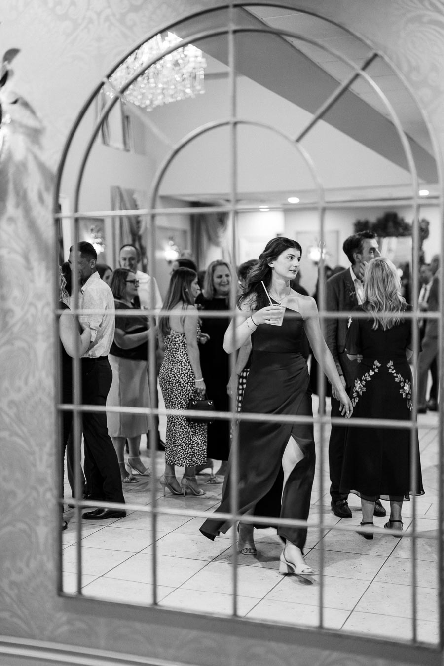 Black and white photo of a woman in a formal dress walking gracefully with a drink, surrounded by a lively crowd reflected in a decorative mirror at an indoor event, featuring elegant decor and chandeliers.