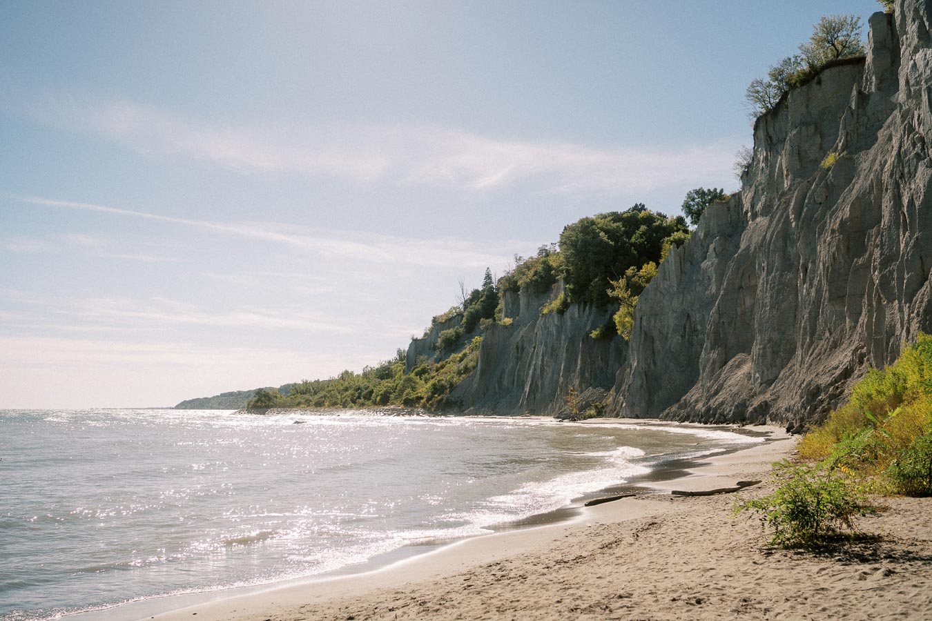 A scenic view of a sandy beach with rugged cliffs and lush greenery, under a clear blue sky, capturing the tranquil shoreline of a coastal landscape.