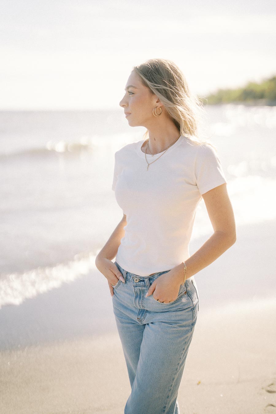Young woman in casual white T-shirt and jeans standing on a sunny beach with gentle waves in the background, looking into the distance.