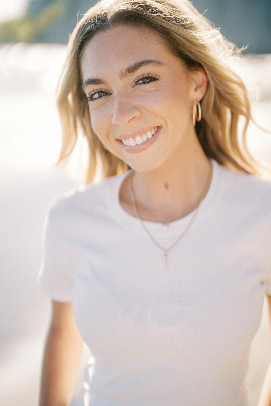 Smiling woman with long hair in a white shirt in a bright outdoor setting.