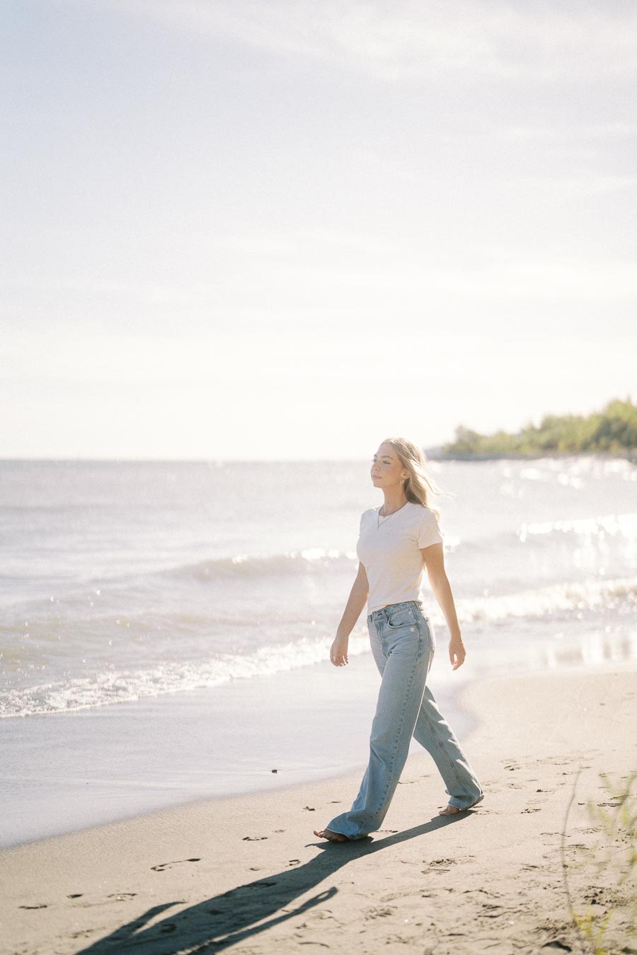 A woman wearing a white t-shirt and jeans walks barefoot along a sunny beach, with gentle waves in the background.