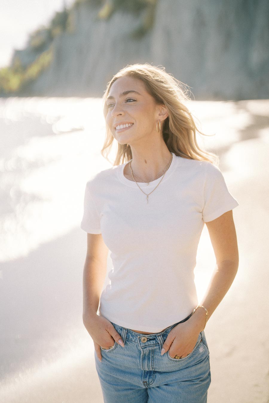 Smiling woman in a white t-shirt and jeans enjoying a sunny beach day, with cliffs in the background.