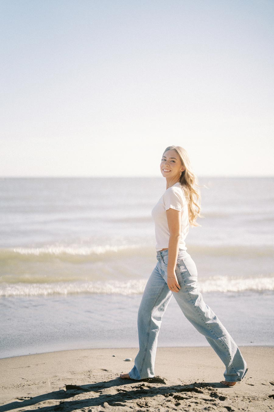 A woman in a casual outfit smiling and walking along the beach, with ocean waves in the background.