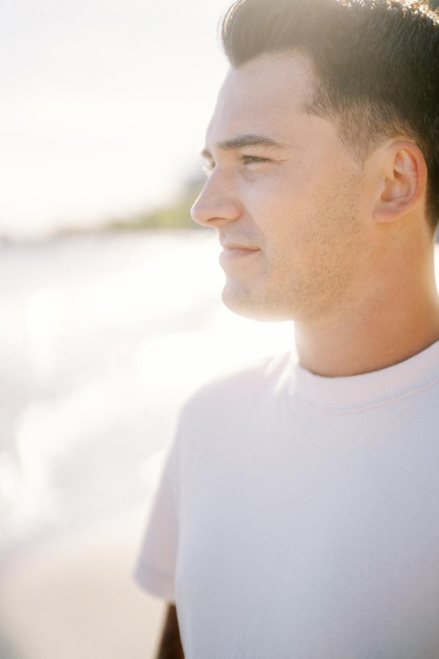A young man in a white shirt stands on a sunny beach, gazing into the distance with a serene expression.
