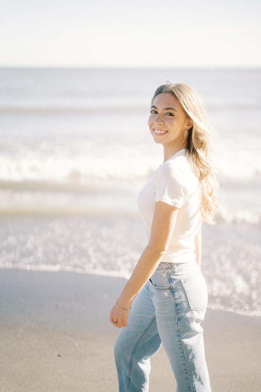 Young woman smiling and walking on a sunny beach, wearing a white t-shirt and blue jeans.