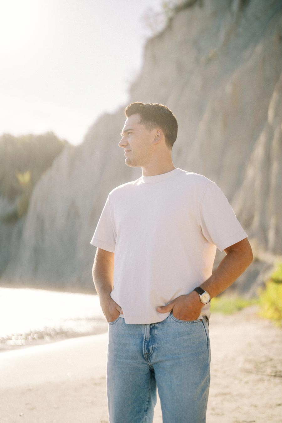 Man standing on a beach with cliffs in the background, wearing a white t-shirt and jeans, enjoying a sunny day.