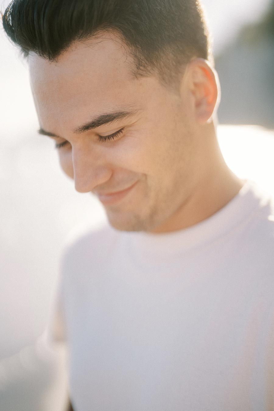 Smiling young man with dark hair, wearing a light shirt, looking down in soft natural light.