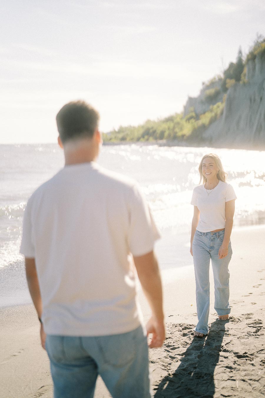 Two people enjoying a sunny day on a beach with gentle waves and lush green cliffs in the background.