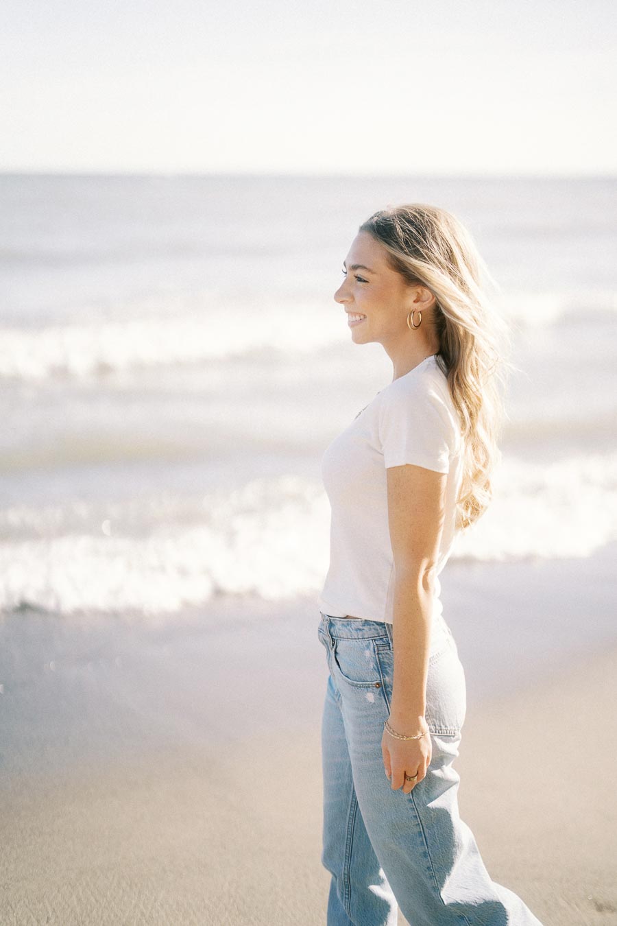A woman in a white t-shirt and jeans enjoys a sunny day at the beach, with gentle waves in the background.