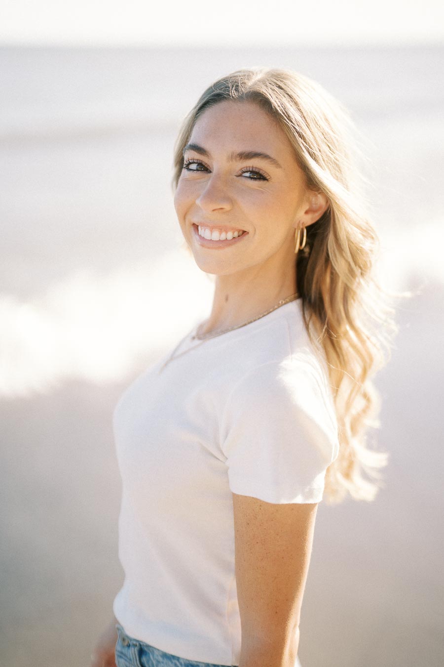 A smiling woman with long blonde hair and hoop earrings, wearing a white t-shirt, stands on a beach with gentle waves in the background, enjoying a sunny day.