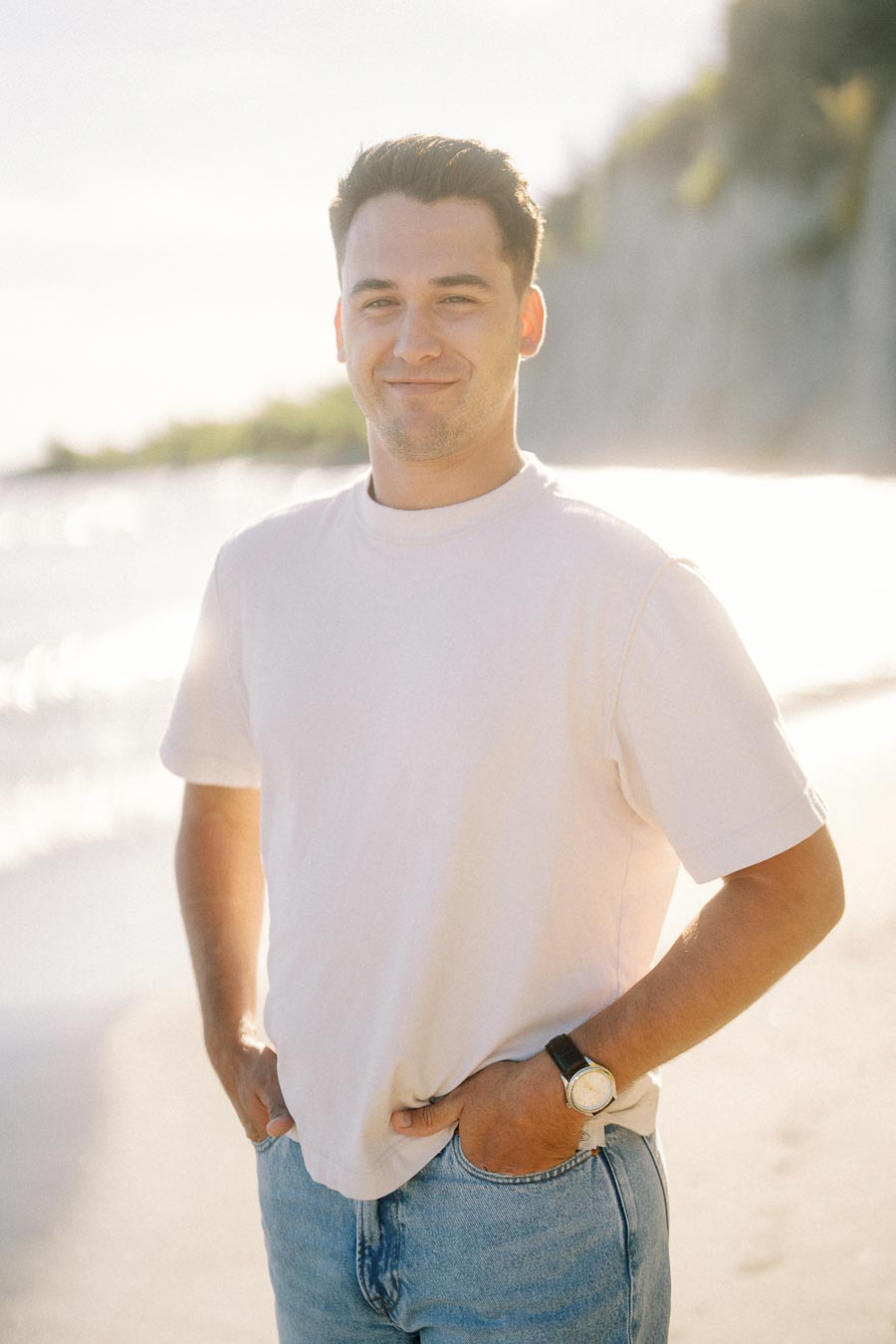 Smiling man in white t-shirt and jeans on a sunny beach with cliffs in the background