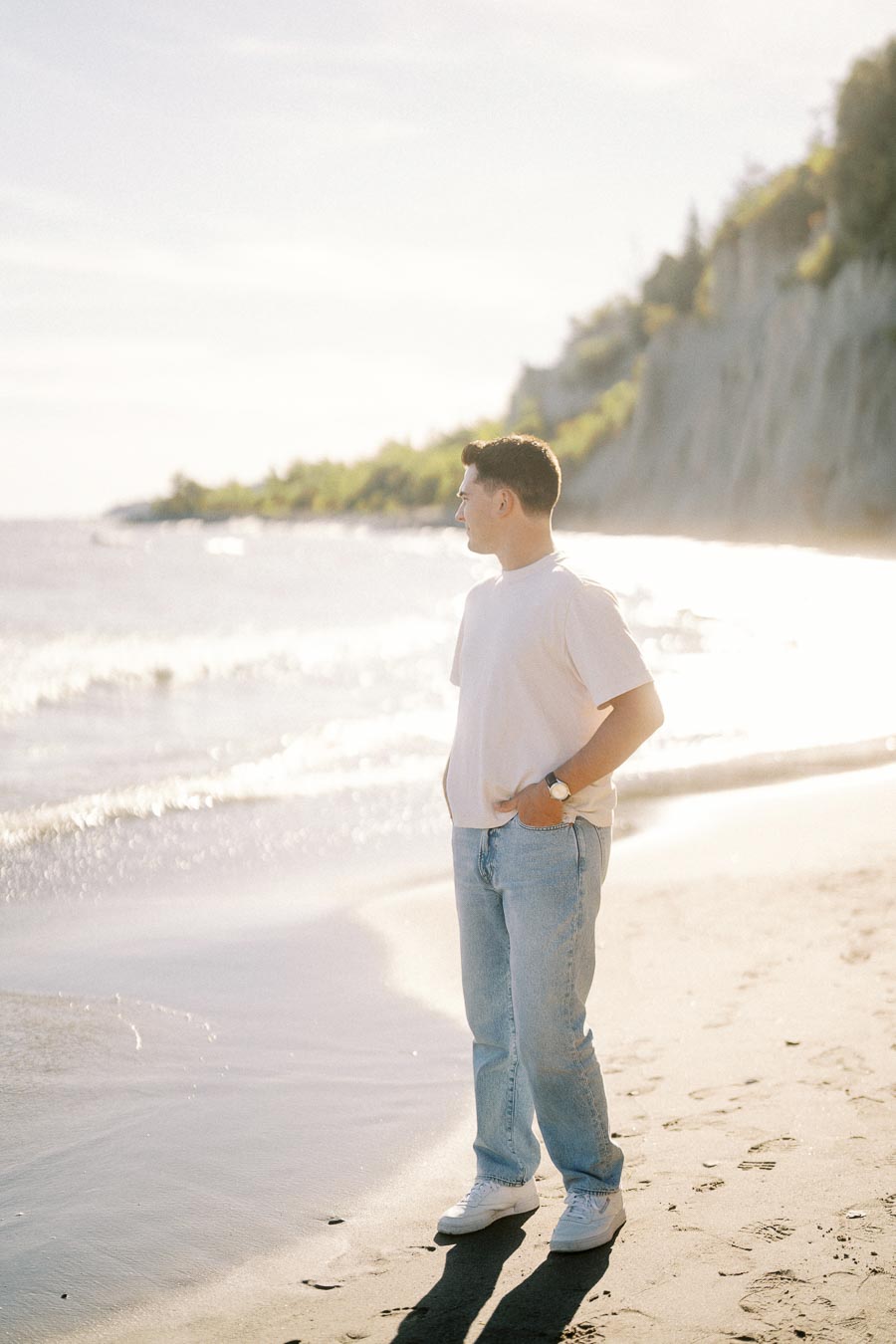 A person stands on a sunlit beach, gazing toward the ocean, with cliffs and greenery in the background, wearing a white t-shirt and jeans.