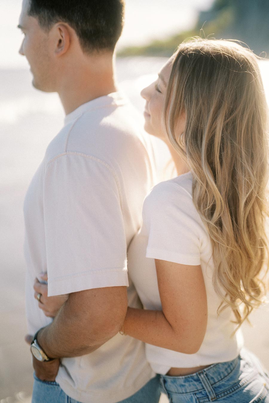 A couple embraces on a sunny beach, wearing casual white shirts and denim, with the ocean and cliffs in the background.