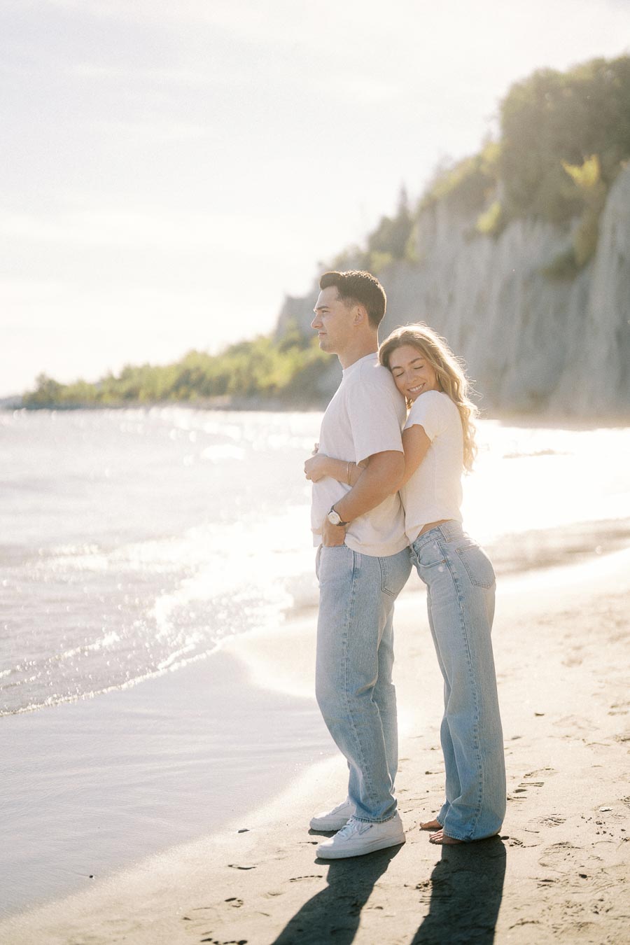 A couple embracing on a sunny beach with cliffs and ocean waves in the background, wearing casual white shirts and jeans.