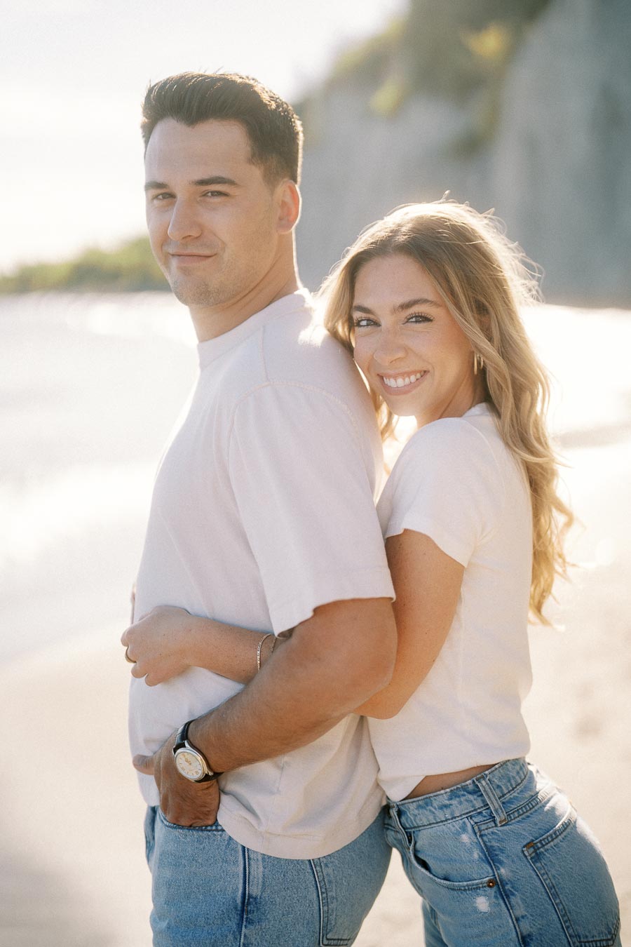 Smiling couple embracing on a sunny beach day, wearing casual white shirts and denim jeans, with scenic cliffs in the background.