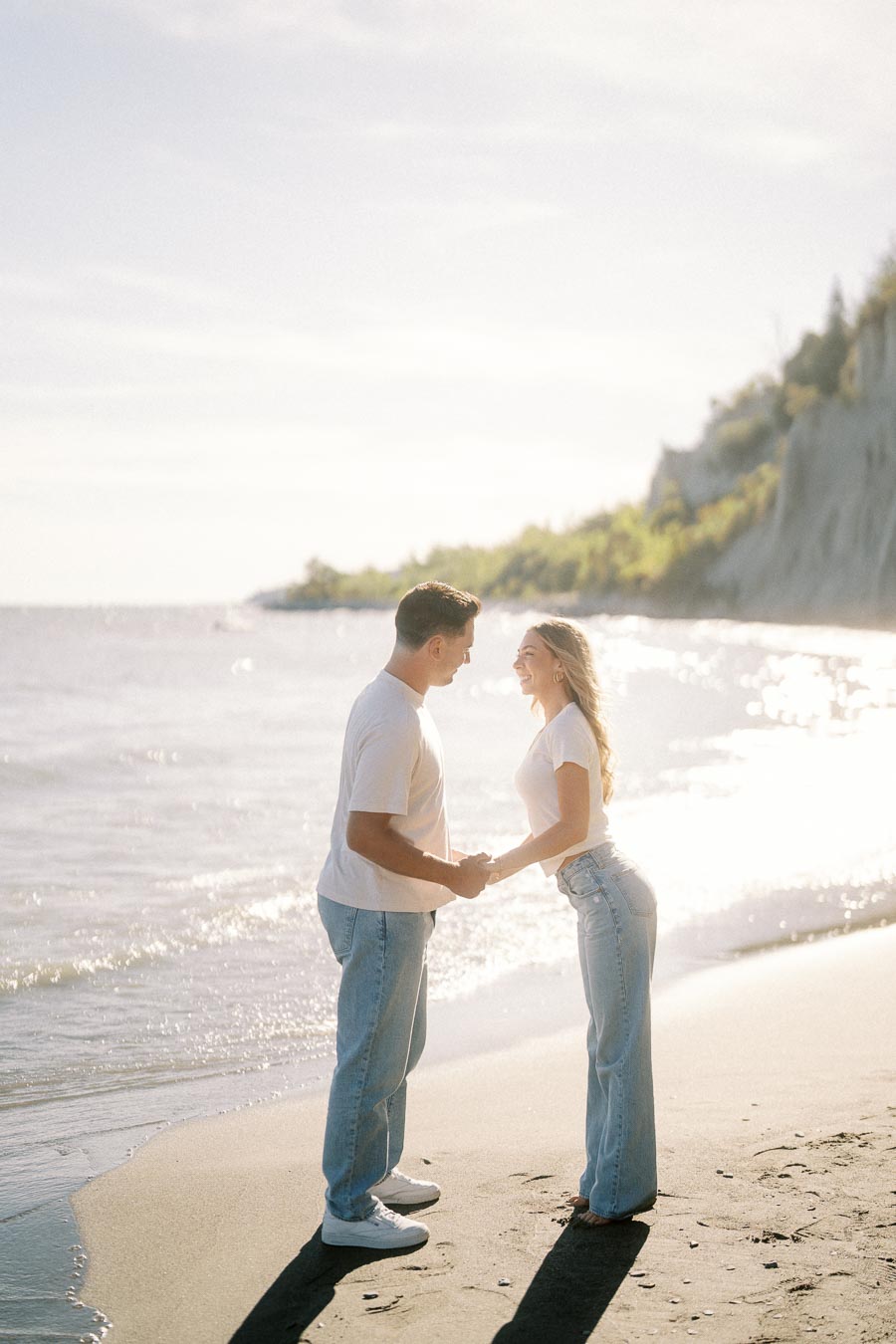 A couple holding hands and smiling at each other on a sunlit beach, wearing casual white tops and jeans, with the ocean and cliffs in the background.