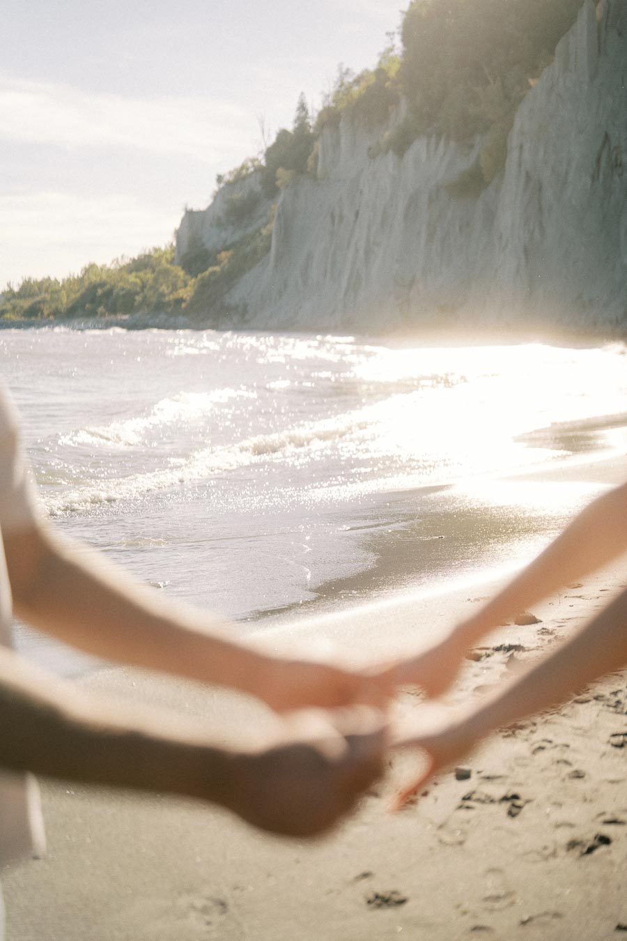 Romantic couple holding hands on a sunny beach with waves lapping the shore and cliffs in the background.