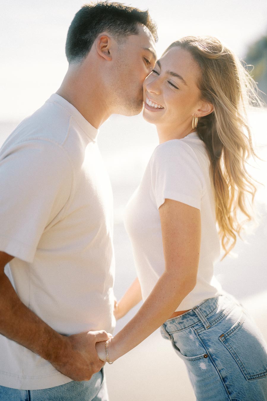 Romantic couple at a beach, man kissing smiling woman on the forehead, both holding hands and wearing casual clothes, sunny day.