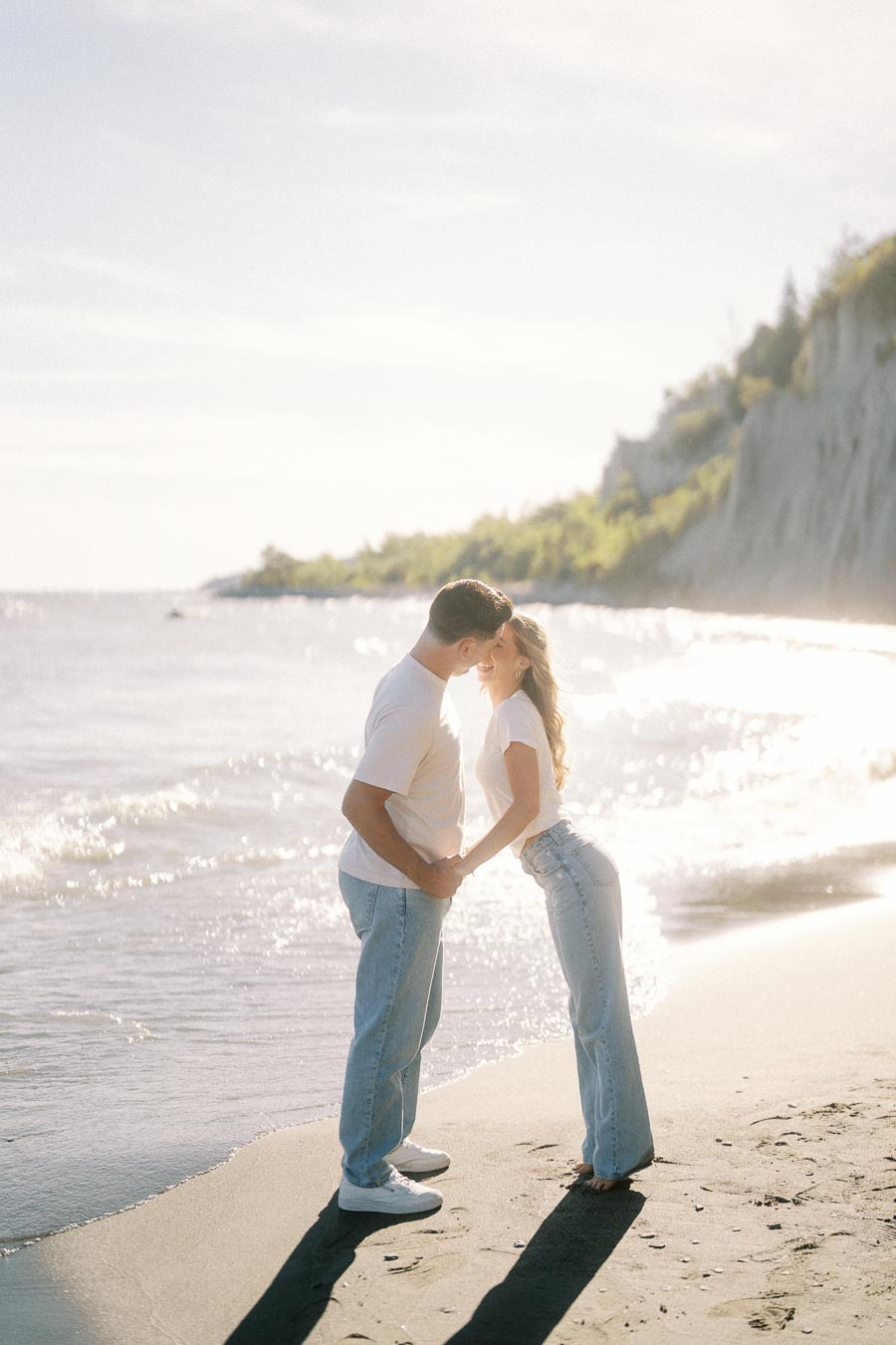 A couple in casual attire shares a romantic moment, kissing on a sandy beach with gentle waves and scenic cliffs in the background, bathed in warm sunlight.
