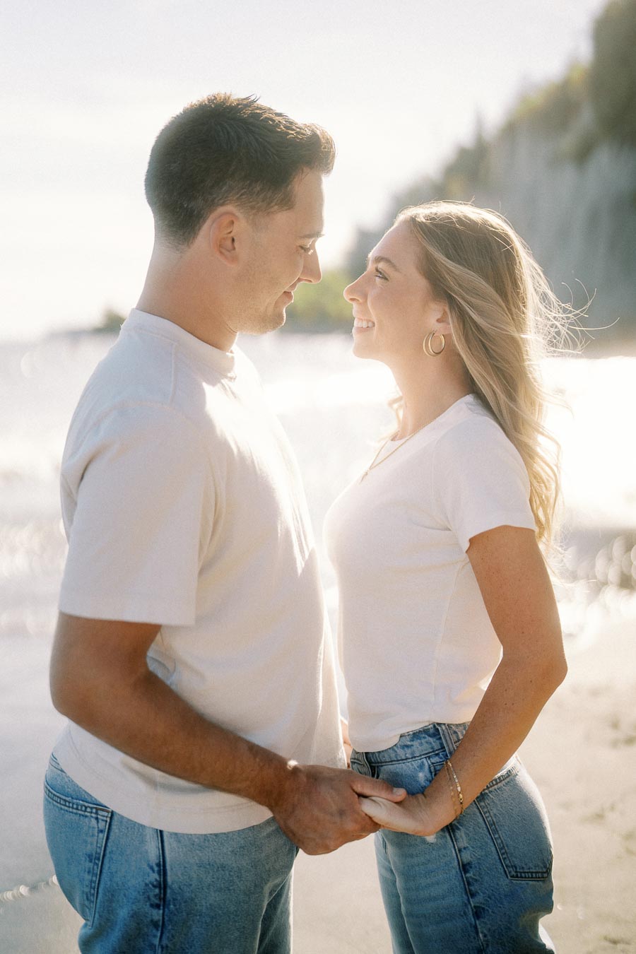 A couple holding hands and smiling at each other on a sunny beach, both wearing casual white shirts and jeans, with the ocean and cliffs in the background.