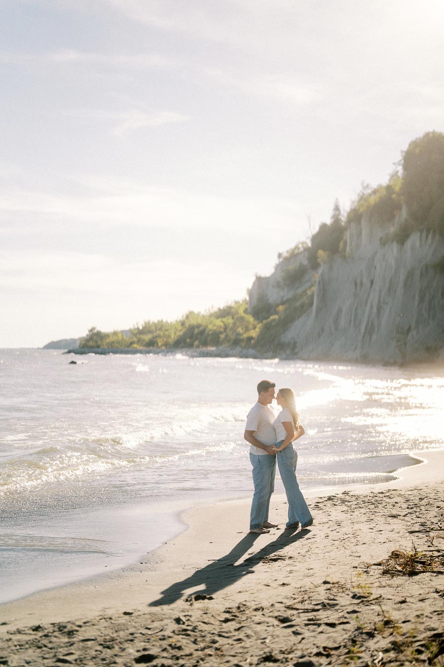 A romantic couple embraces on a sunlit beach with cliffs and sparkling ocean waves in the background, capturing a perfect seaside moment.