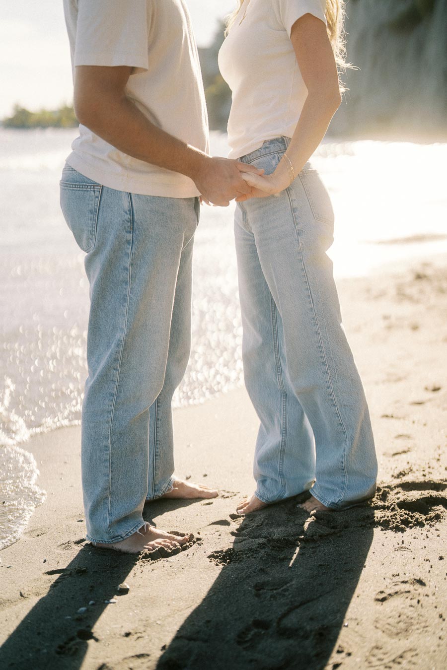A couple holding hands on a sunny beach, wearing casual jeans and t-shirts, with waves gently washing against the sandy shore.