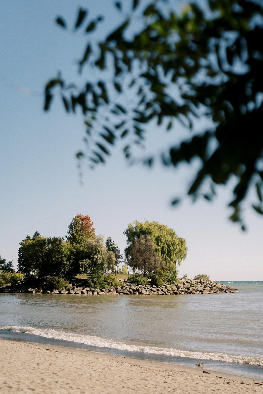 Scenic view of a sandy beach with gentle waves, leading to a small, lush green island with various trees, under a clear blue sky.