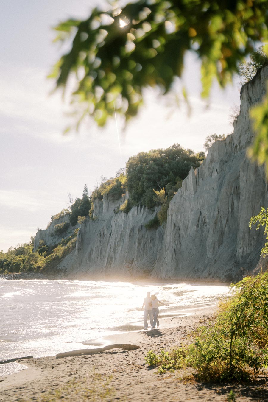 Scenic beach view with a couple walking along the shoreline, surrounded by sunlit cliffs and lush greenery, under a bright sky.