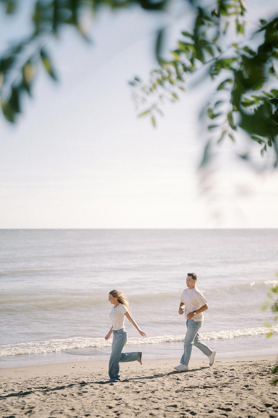 A couple running joyfully along a sandy beach with gentle waves in the background, framed by leafy branches under a clear blue sky.