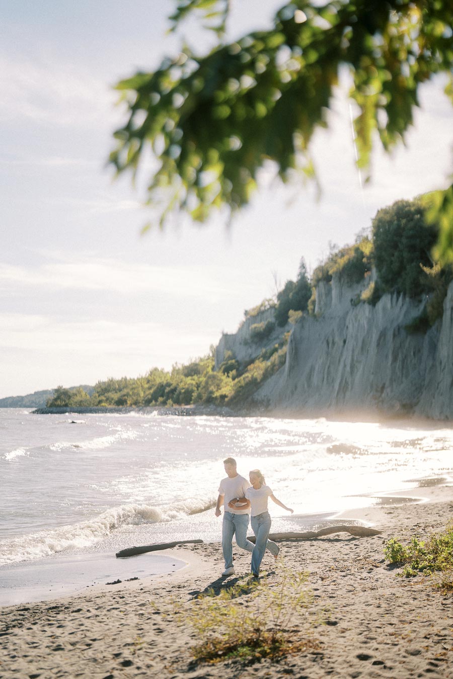 A couple walking along a sunny beach, with waves gently hitting the shore and lush green cliffs in the background.