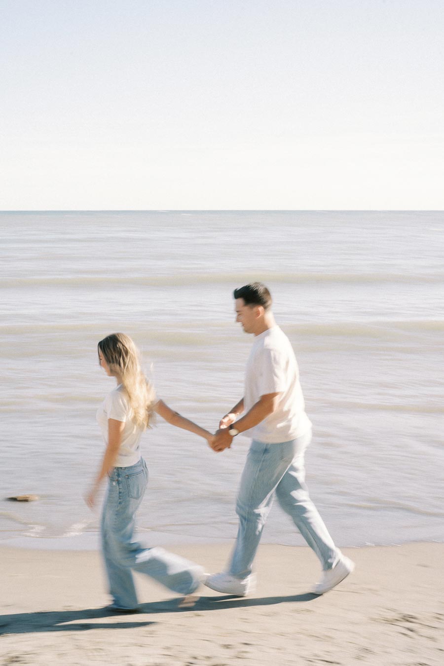 A couple holding hands and walking along a sandy beach with gentle waves in the background on a sunny day.