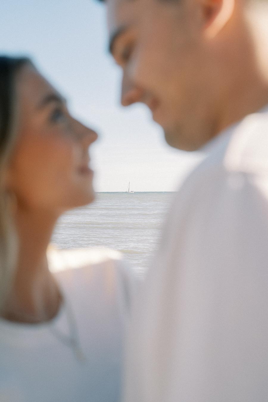Blurred close-up of two people looking at each other with a serene ocean and distant sailboat in the background, conveying a romantic and tranquil beach setting.