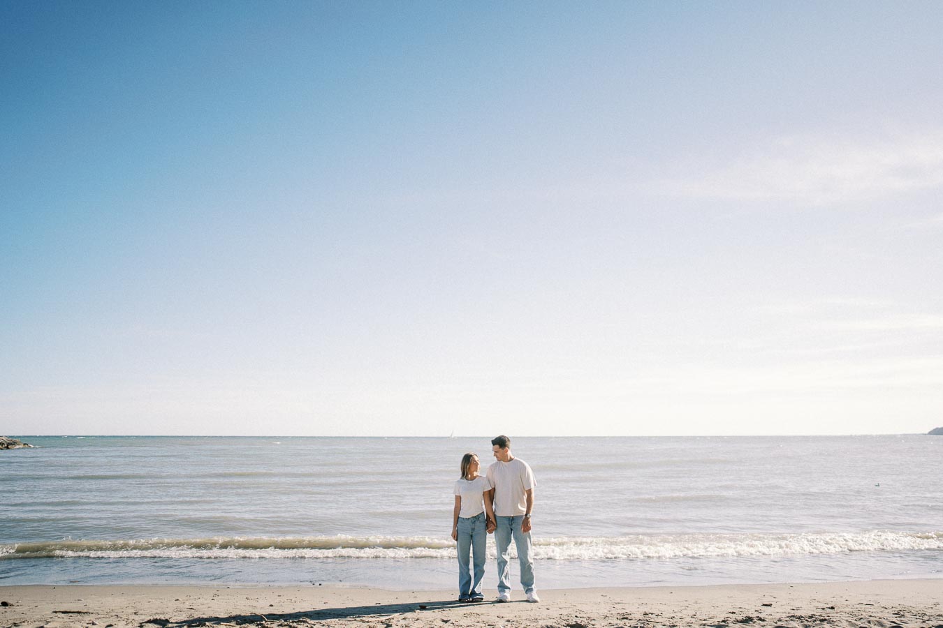 A couple holding hands and standing on a sandy beach with gentle waves and a clear blue sky in the background.