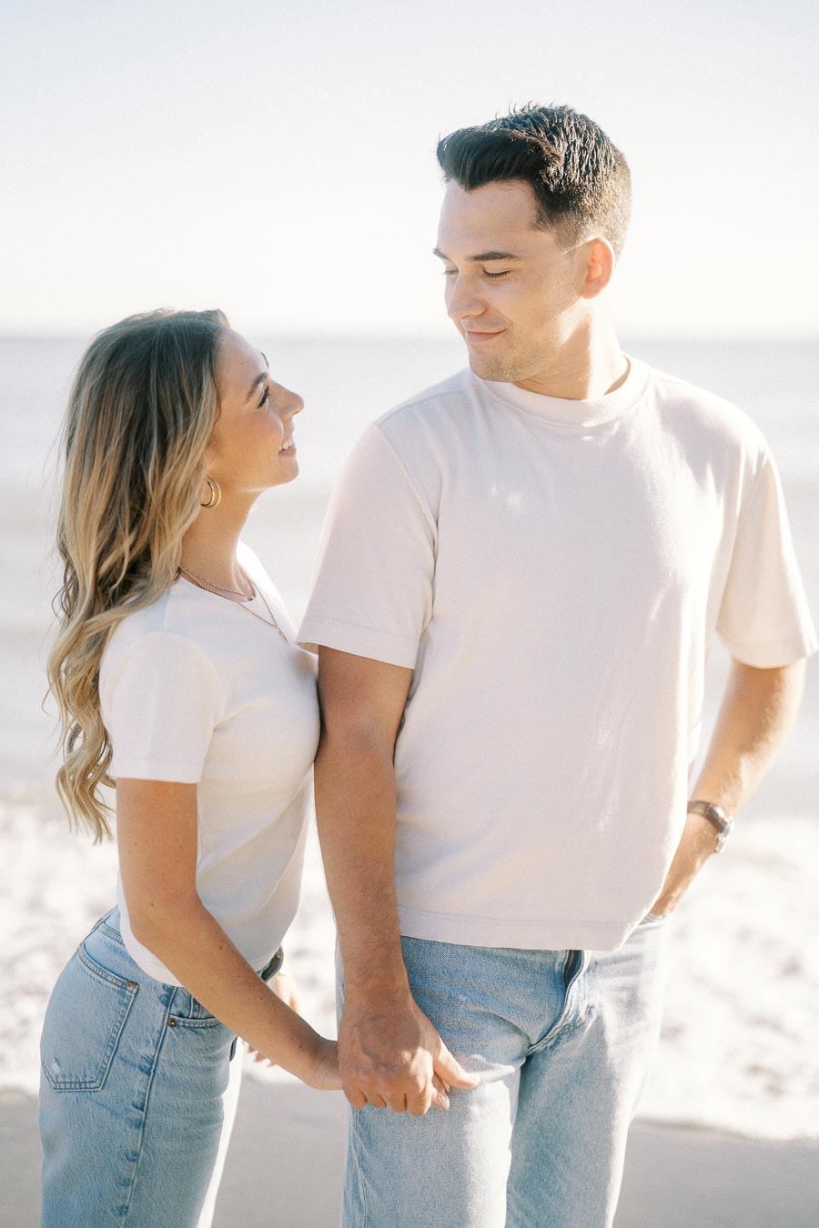 A couple holding hands and smiling at each other on a sunny beach, both wearing casual white shirts and jeans, with the ocean in the background.