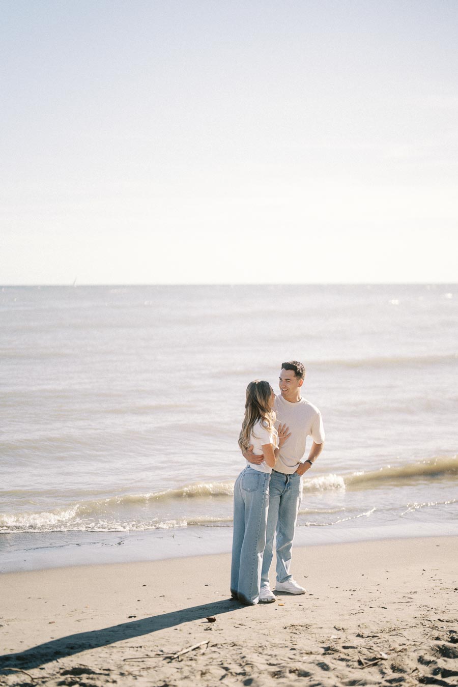 Couple in casual attire embracing at the beach with gentle waves in the background under a clear sky.