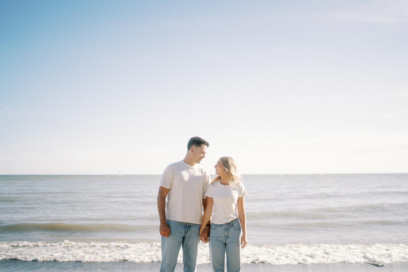 A couple holding hands and smiling at each other while standing by the ocean on a sunny day, wearing matching white shirts and jeans.