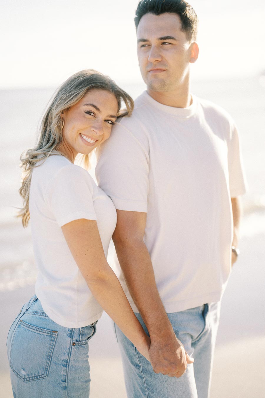 A smiling couple holding hands on a sunny beach, both wearing casual white shirts and blue jeans, with an ocean backdrop.