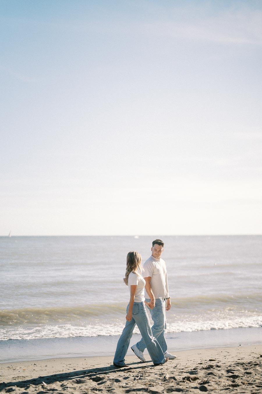 A couple walking hand in hand along a sandy beach with ocean waves in the background, wearing casual clothes and smiling under a clear blue sky.