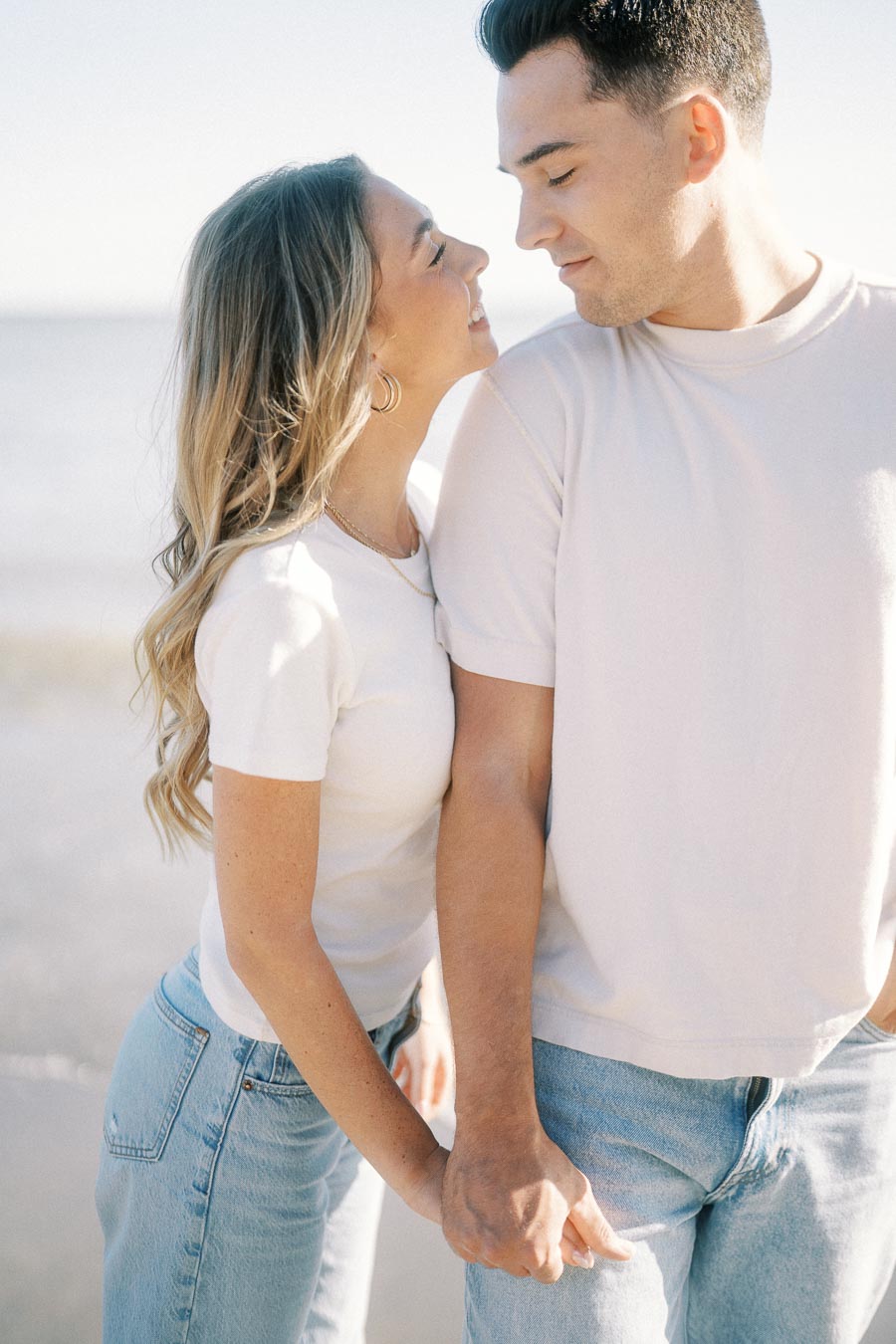 Young couple in casual outfits smiling and holding hands by the beach on a sunny day.