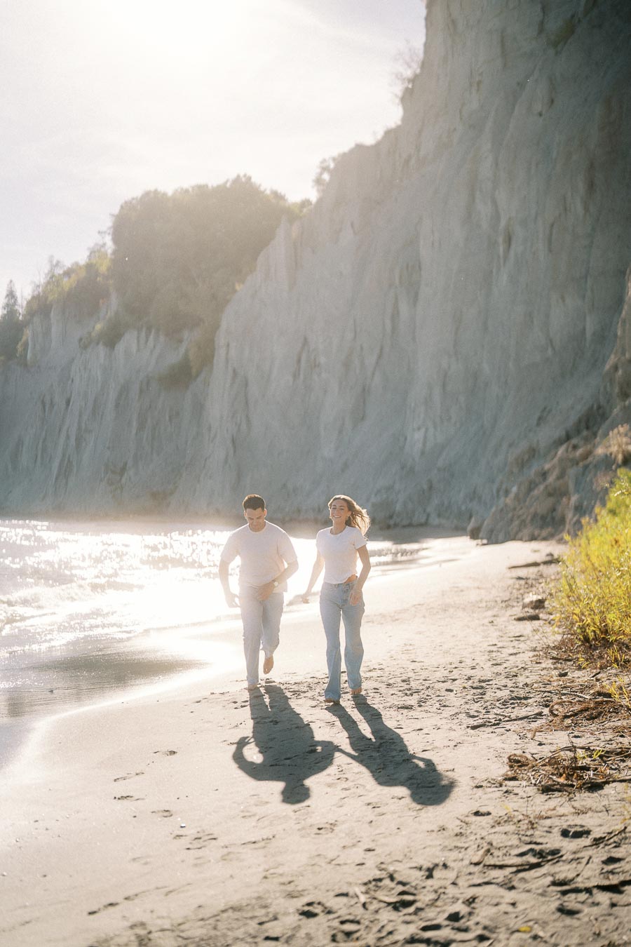 A couple running along a sunlit beach beside towering cliffs, with clear sky and ocean waves in the background.