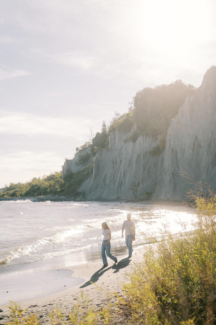 Couple walking along a sunny beach with cliffs in the background, surrounded by lush greenery and the gentle waves of the ocean.
